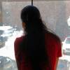 A woman with long brown hair, wearing a red T-shirt, looks out a window into a parking lot. You cannot see her face.
