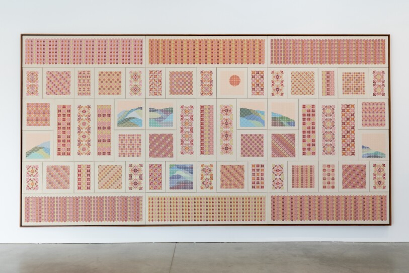 Hanging on a gallery wall is a large scale tapestry, featuring a ranger of geographic shapes, floral patterns and patterns evoking mountain scapes, the colors are primarily yellows, pinks, purples, with splashes of green and blue