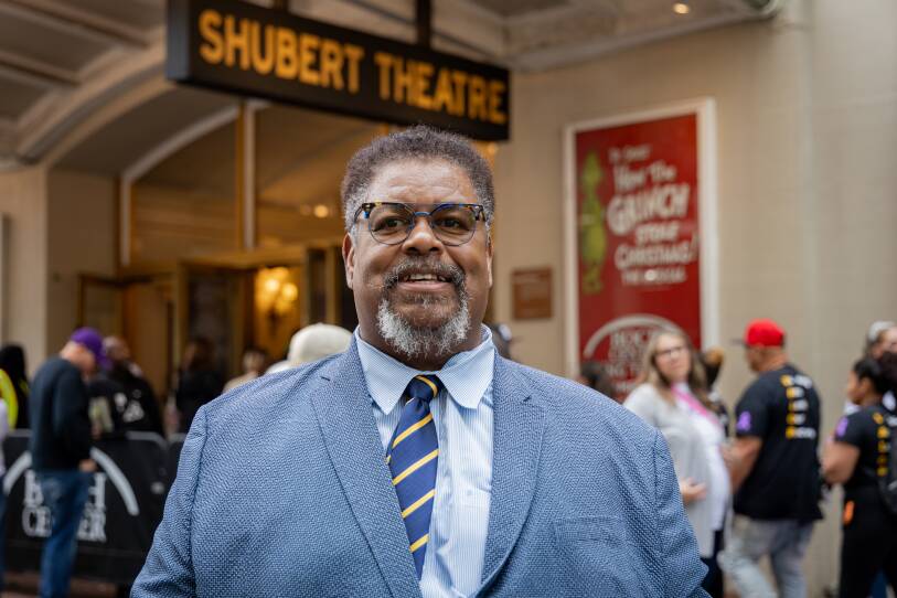 A man in a suit and tie smiles slightly as he stands on the sidewalk by a theater.