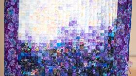 This is a photograph and a patchwork quilt. The pattern is made of an intricate series of fabric squares.  The middle of the quilt is a gradient, with darker colors fragmenting into lighter ones as you move your eyes up the quilt. The border is purple and the pattern in floral.