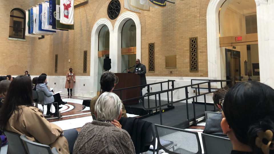 A man speaks at a podium in front of dozens of audience members.