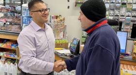 A white man with black rimmed glasses smiles as he shakes the hand of a white man in a blue jacket and black beanie hat.