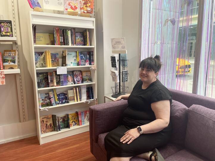 A woman wearing a black jersey dress and black sandals sits on a purple sofa. Behind her is a bookcase lined with books. A large window, that offers a view of the street,  is behind her.