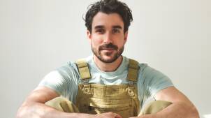AJR's Adam Met sits on the floor. He is wearing brown overalls and a teal shirt.
