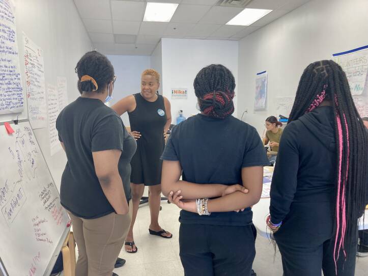 Three teenage Black girls face away from the camera, listening to and looking at a Black woman surrounded by whiteboards and lists on long sheets of paper.