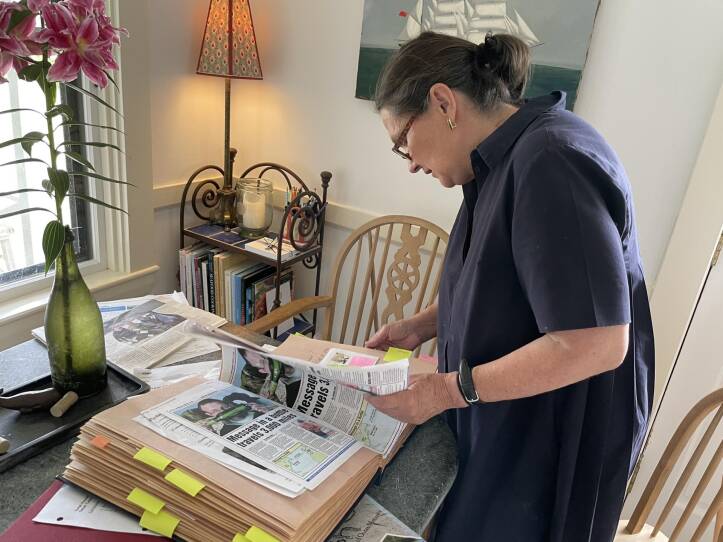 A woman stands at a dinning room table, flipping through a scrapbook full of newspaper clippings.