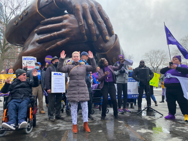 A crowd of people, many wearing purple and some in wheelchairs, stand in front of the Embrace statue and wave their hands. One person holds a sign that says 'integration not institutionalization.'