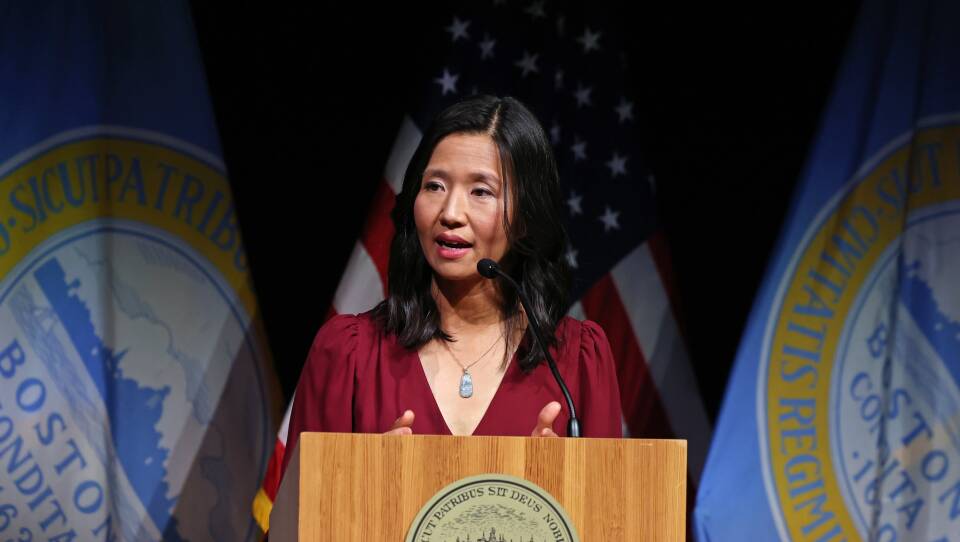 Michelle Wu speaks from behind a lectern on stage, with Boston city flags hanging behind her.