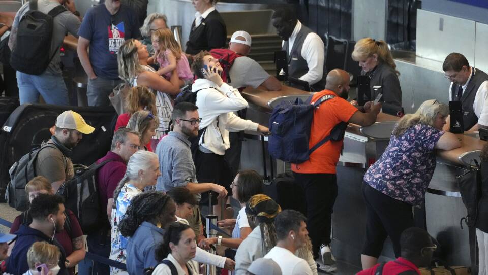 A crowd of people with luggage look upset and frustrated while waiting at an airport service counter.