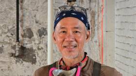 A man stands inside a building under construction. He is wearing a bandana and apron, and has a respirator around his neck.