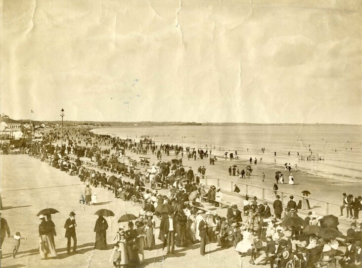 A sepia photograph of a beach with people wearing long dresses, suits and holding umbrellas to protect from the sun.