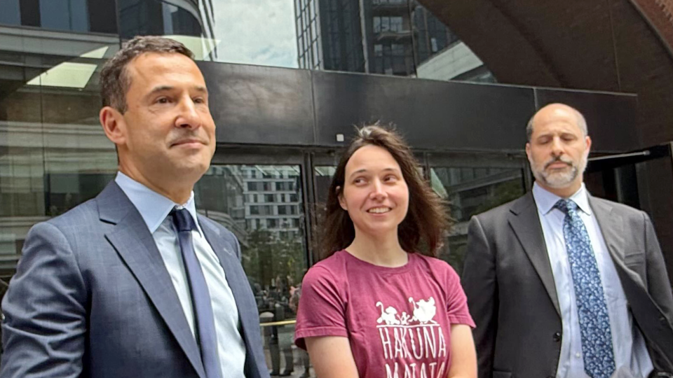 A woman in a T-shirt is flanked by two men in suits. She's smiling, standing outside a courthouse.