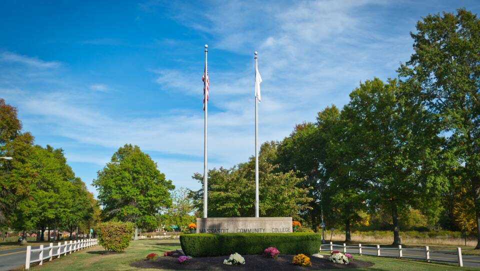 Two flagpoles stand behind a gray sign that reads "Massasoit Community College," with white, pink and orange flowers in the landscaping nearby.