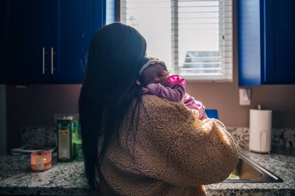 A mother and her newborn stand at the kitchen sink.
