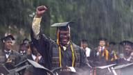 A young black man in a graduation gown holds his fist up in celebration as he's surrounded by other grads.