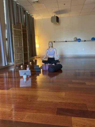 A woman is seated on a yoga mat with her eyes closed and her hands out at her sides.