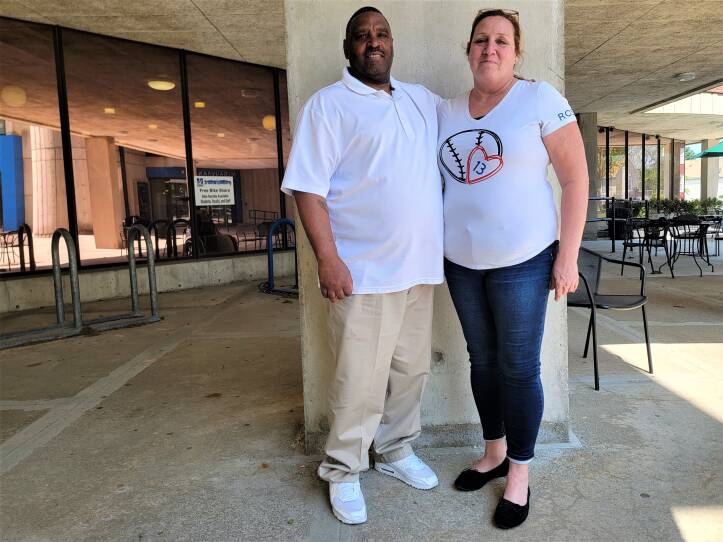 A couple stands together outside, with their arms around each other and smiling for the camera.