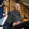 A woman in a business-y black-and-white patterned jacket holds a gavel as she stands behind a speaker's podium in the Massachusetts Statehouse.