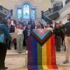 Several people stand on a staircase in the State House with their right hands raised. A woman on the left reads from a piece of paper and a sign language interpreter is on the right. In the middle, a pride flag is draped over a podium.