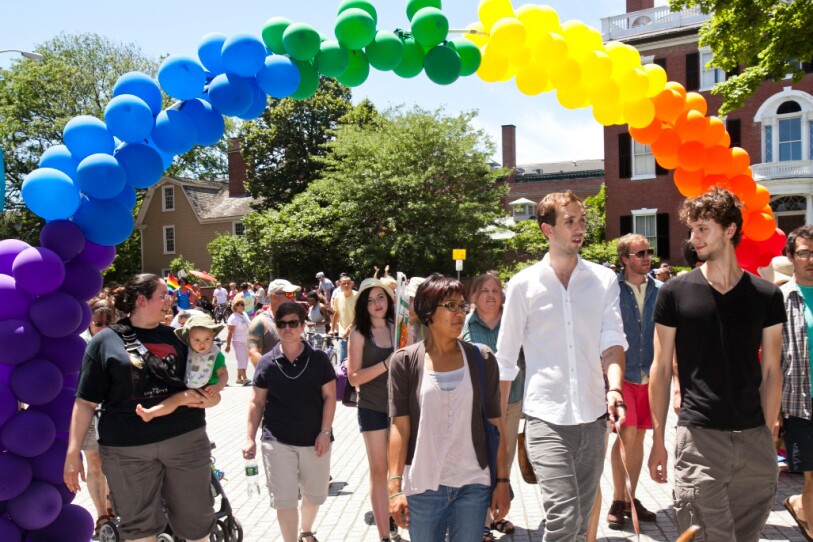 Dozens of people walk through a New England town, passing under a rainbow balloon arch.