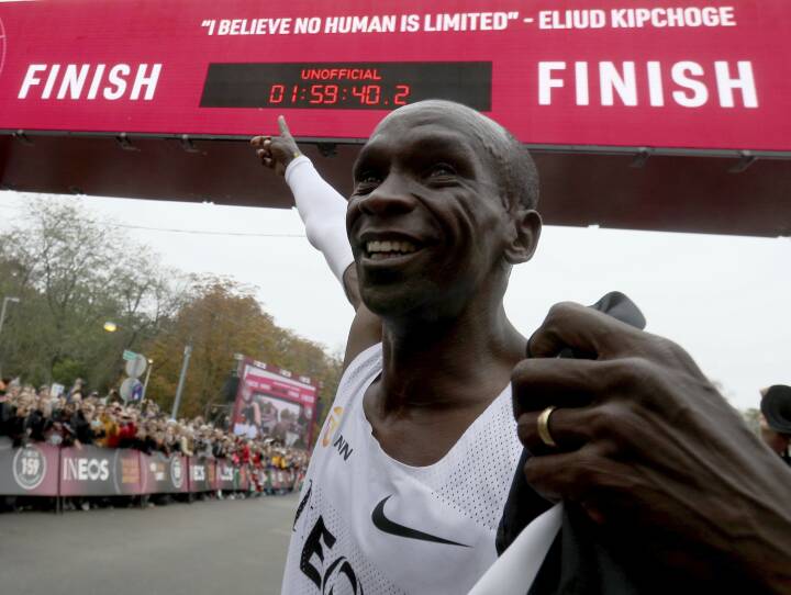 A man beams with a smile as he points to the digital clock at a race finish line. The clock reads 1 hour, 59 minutes and 40 seconds.