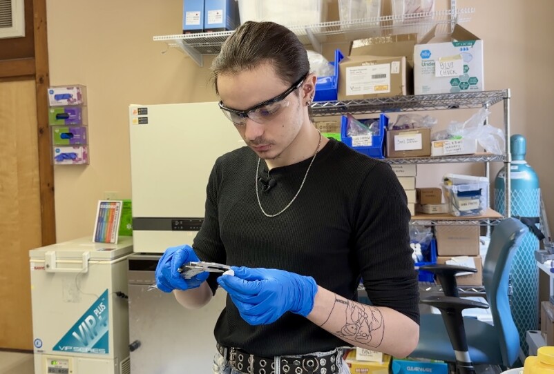 A young white man in a lab wearing safety goggles a black shirt and blue rubber gloves and holds a caliper to measure a small object.