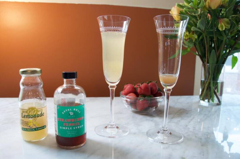 Strawberry-fennel, and lemonade syrup bottles, in front of Mimosa glasses.