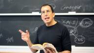 A man wearing a black shirt and glasses holds a book in front of a chalkboard and gestures with a pointed finger.
