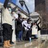 A small group of people wearing winter jackets and boots raise their arms in the air and hold signs as they stand at the top of a small staircase leading to the gate of the Massachusetts State House.
