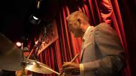 Joe Farnsworth sits at a drum kit in front of a red curtain.