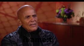 This is a photograph of Harry Belafonte. He's sitting sown. He's wearing a black, down jacket. behind him is a credenza with a vase of flowers on it.