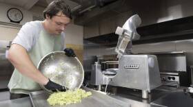 A person wearing gloves and a hair net is seen emptying a bowl of chopped vegetables into a tray.