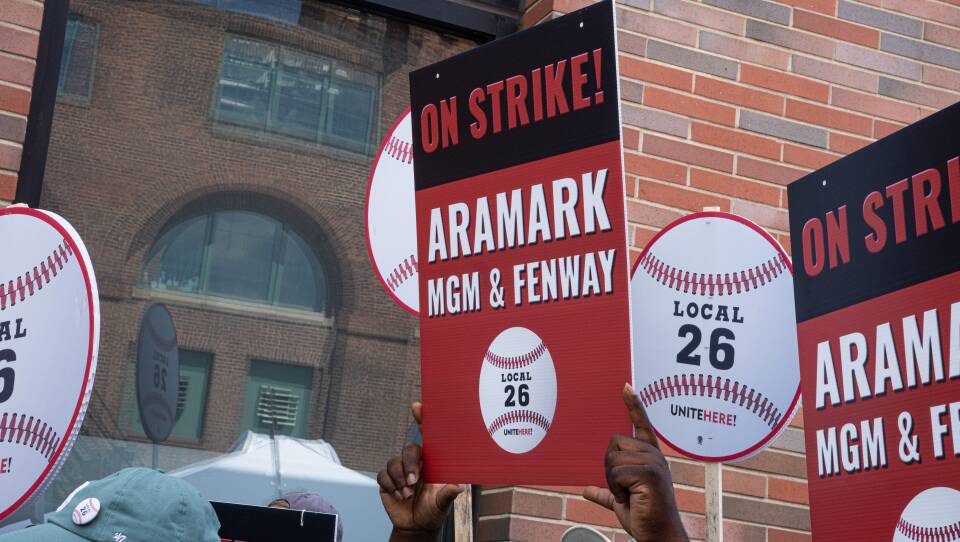 Hands hold up a red, black and write sign that reads: On Strike, Aramark MGM & Fenway, Local 26.