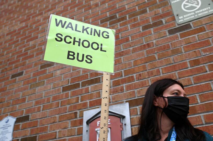 A woman wearing a mask outside a brick building holds a yardstick that has a laminated neon green sign at the end, reading "walking school bus"