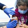 A nurse wearing gloves places a bandage on the arm of a young girl as she sits on her father's lap.
