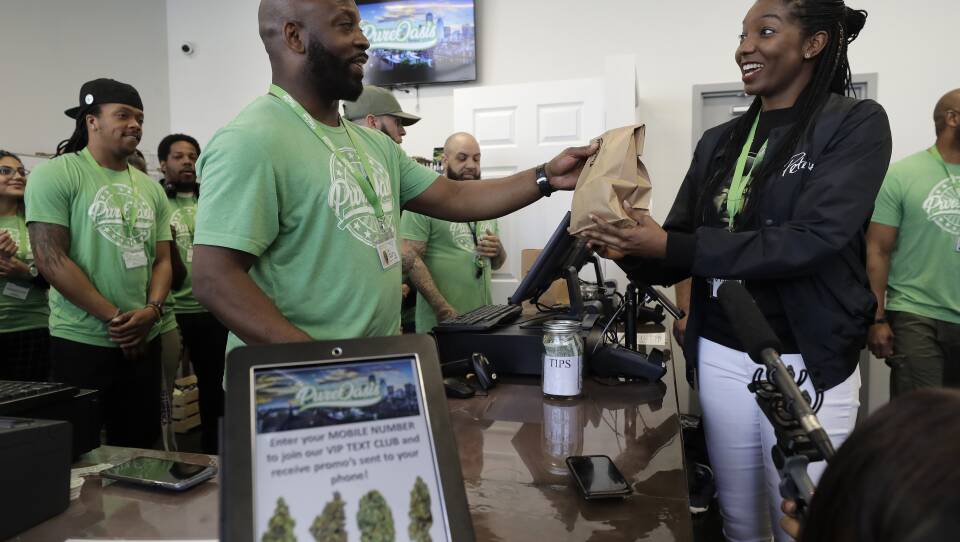 A man behind a counter and register hands a brown paper bag to a woman. Other people stand around watching.