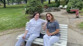 Two women sit on a bench in a grassy area, with a fountain nearby.