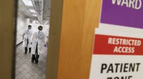 Two health care workers are photographed through a door's window, wearing protective gear