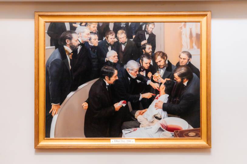 A large painting in a golden frame shows about 20 men dressed in black, period clothing looking on as one man operates on an unconsicous patient, whose neck is bloody. One figure holds a glass jar of ether near the unconsious man's face.