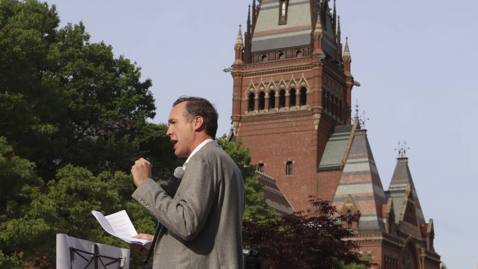 A man in a suit holds a piece of paper with notes as he speaks into a microphone on Harvard's campus.