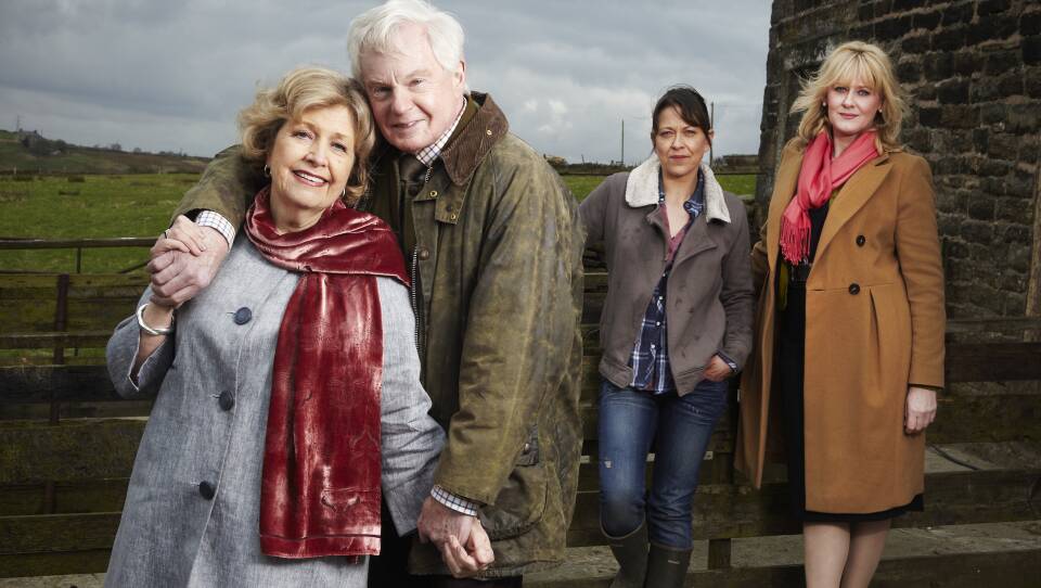 An older couple stands near the camera, embracing and smiling at the viewer. Behind them, two adult women, their daughters, stand together, but apart, giving smaller smiles to the viewer.
