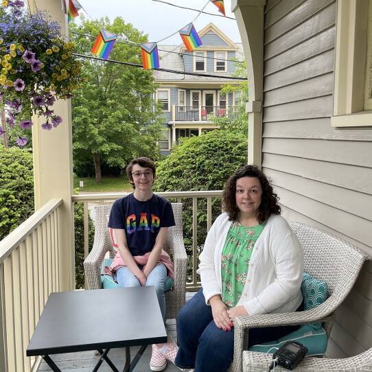 A teenager wearing glasses and a GAP t-shirt with rainbow lettering sits in a wicker chair on a home's porch next to their mother, who has a cardigan and is holding her hands in her lap.