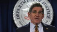 A man in a suit speaks in front of a Justice Department seal.