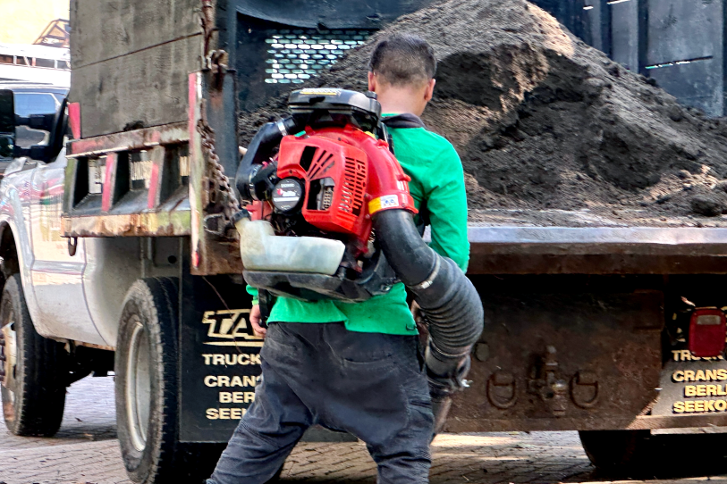A man in a T-shirt and pants using a leaf-blowing machine near a landscaping truck