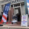 A fabric pop-up banner in front of a large government building says "vote here". A second sandwich board says "early voting here".