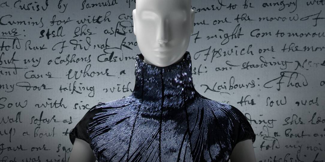 A mannequin wears a dark glittery high-neck dress against a white wall with black cursive writing.