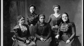 Five women dressed in finely tailored black dresses from a bygone era pose and look toward the camera in a black-and-white photograph.