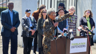 A woman in a green coat points while speaking in front of a group of people at a podium with a sign that reads "Franklin Park Defenders."