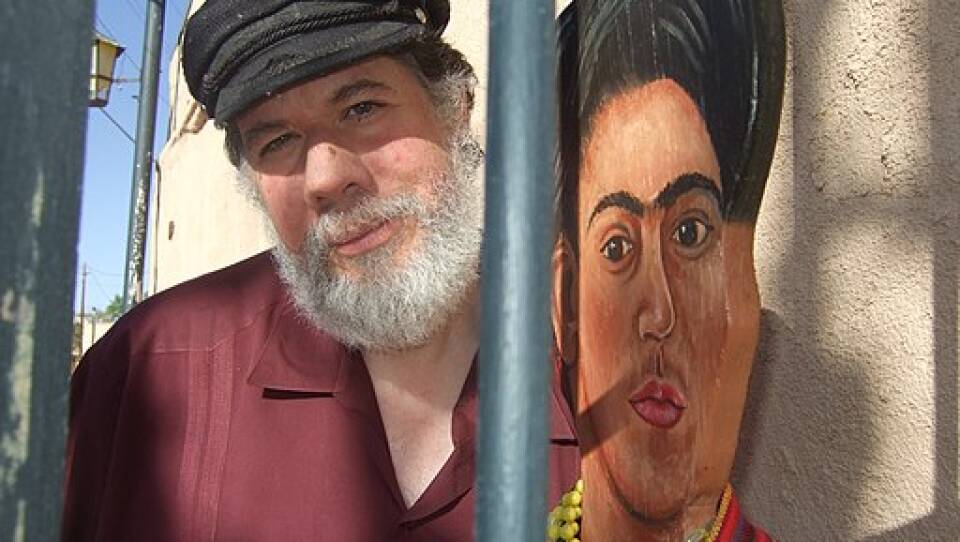 A man with a white beard and black hat stands next to a wooden statue of the artist Frida Kahlo.