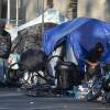 Several tents covered in tarps line the sidewalk. Two men in sweatshirts are standing and talking, and another woman stands nearby smoking a cigarette.